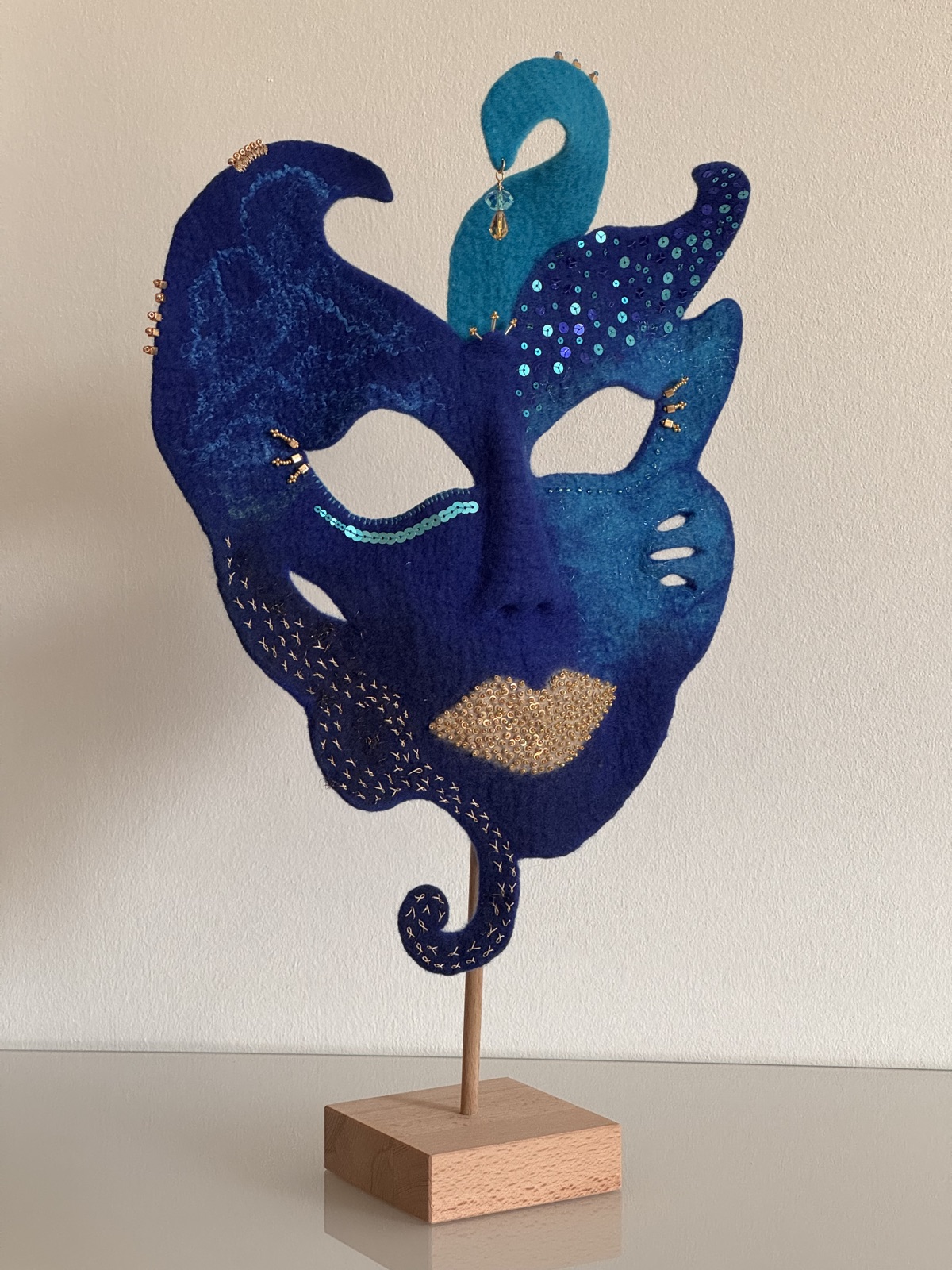 Handgefilzte Maske „Zwischen Traum und Tiefe“ in Blau und Türkis mit Pailletten und golden bestickten Lippen, auf einem Ständer aus Buchenholz